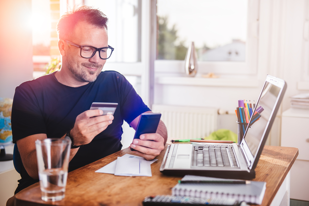 Um home está em sua mesa de trabalho,, ele usa óculos e veste uma camisa azul. Ele segura um celular e um cartão de crédito, procurando entender como funciona o cashback do cartão scale. na mesa também há papéis com anotações e um notebook